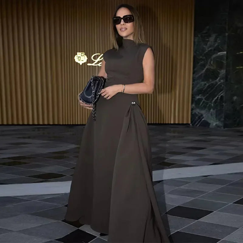 Woman in a brown dress holding a handbag in an indoor setting with a gold logo in the corner.