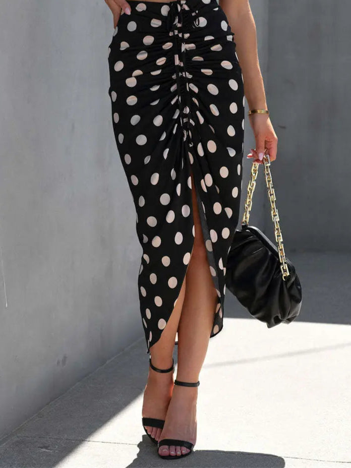 Person wearing a black polka dot skirt with a high slit, holding a black handbag against a gray wall.