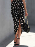Person wearing a black polka dot skirt with a high slit, holding a black handbag against a gray wall.