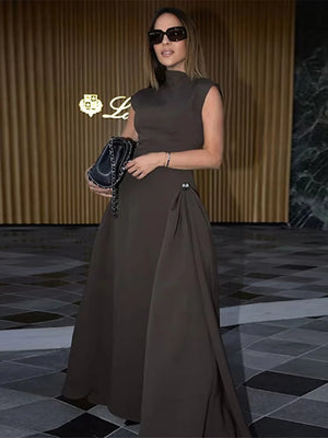 Woman in a brown dress holding a handbag in an indoor setting with a gold logo in the corner.