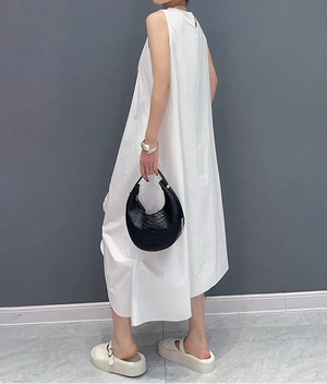 Woman wearing a white sleeveless dress holding a black handbag against a gray wall.