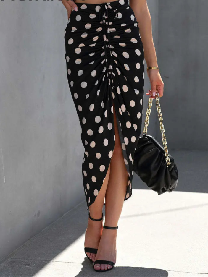Person wearing a black polka dot skirt with a high slit, holding a black handbag against a gray wall.