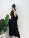 Person wearing a black sleeveless top and ruffled black skirt standing against a white wall with green plants.