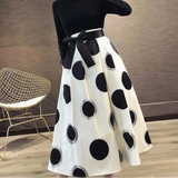 Person wearing a black turtleneck and white skirt with black polka dots, standing indoors.