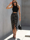 Woman wearing a black top and polka dot skirt with sunglasses and a handbag, standing against a gray wall.